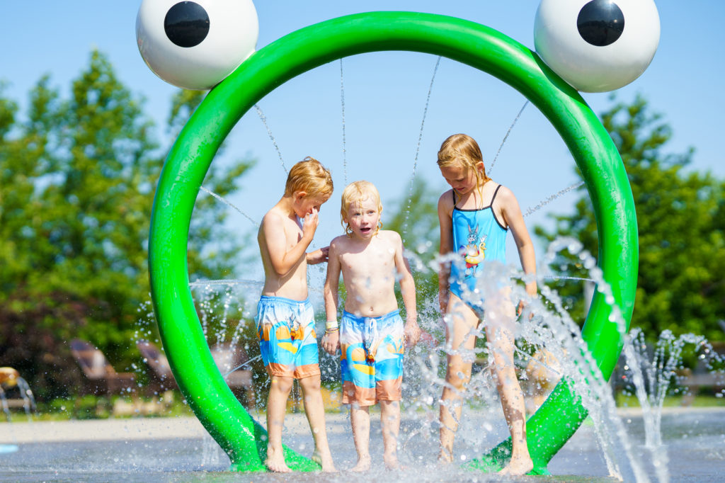 Tre barn som står under en grønn bøyle med froskeøyne og vann som spruter. Foto